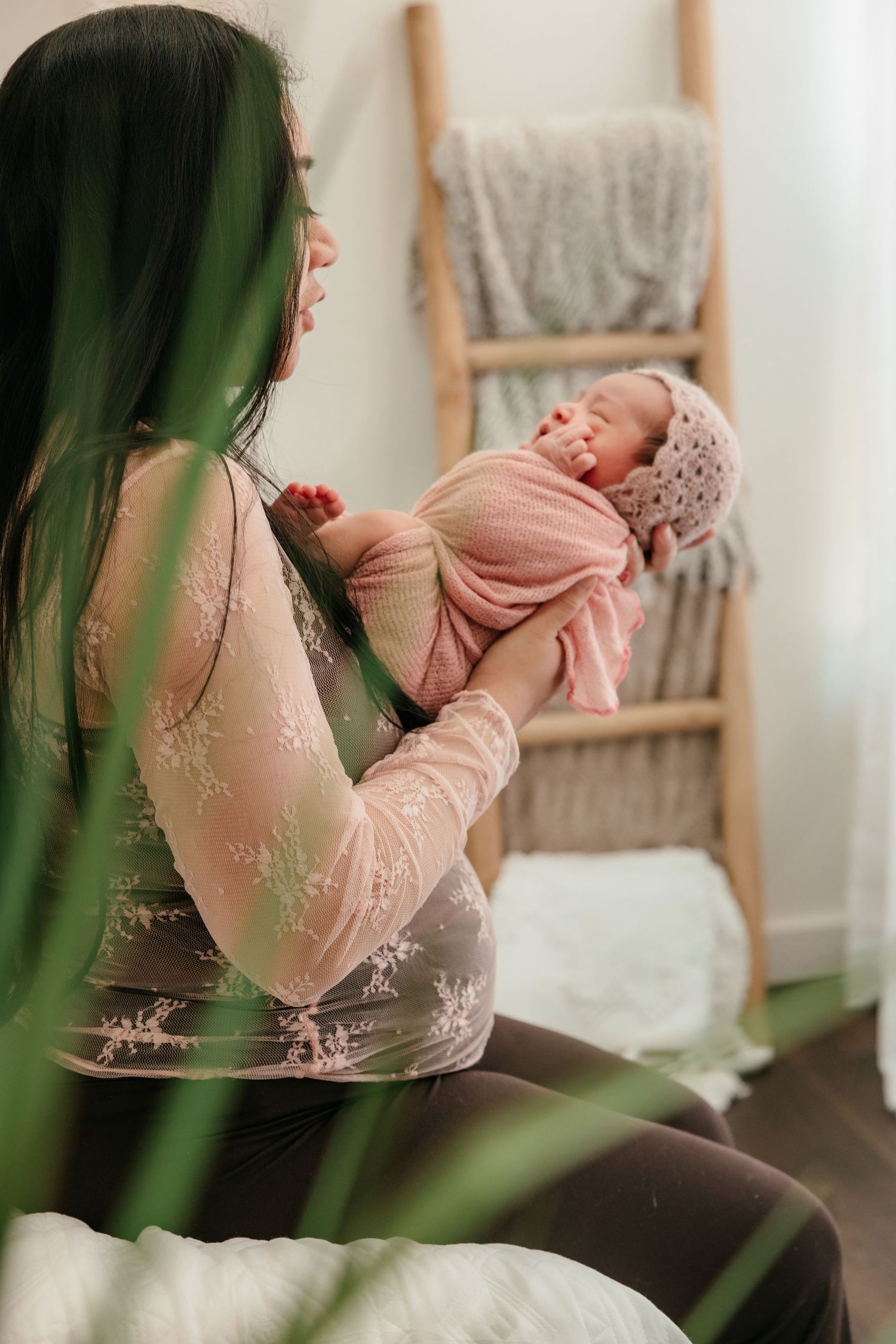 A mother tenderly holds her sleeping newborn wrapped in pink, seated near a decorative wooden ladder with blankets.