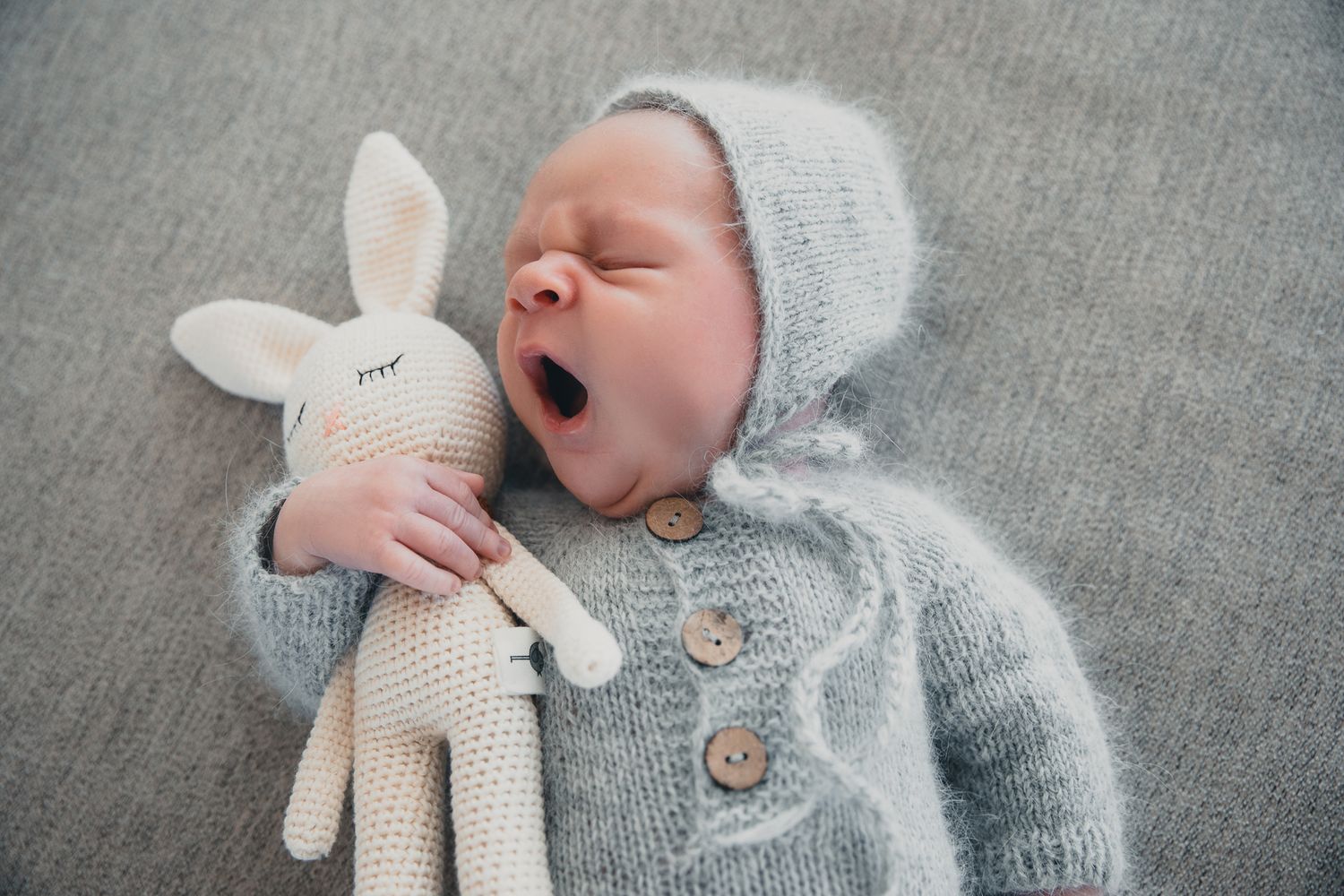 Newborn baby yawning in a gray knit outfit and bonnet, holding a crocheted bunny toy while sleeping peacefully.