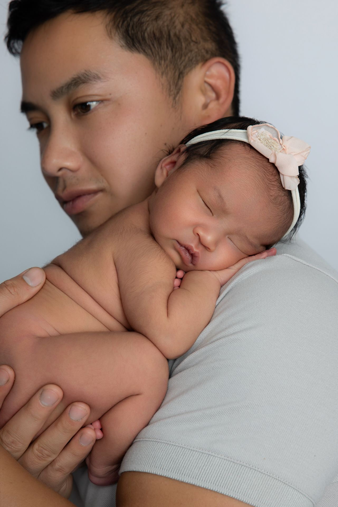 A father tenderly holds his sleeping newborn baby girl wearing a floral headband against his chest.