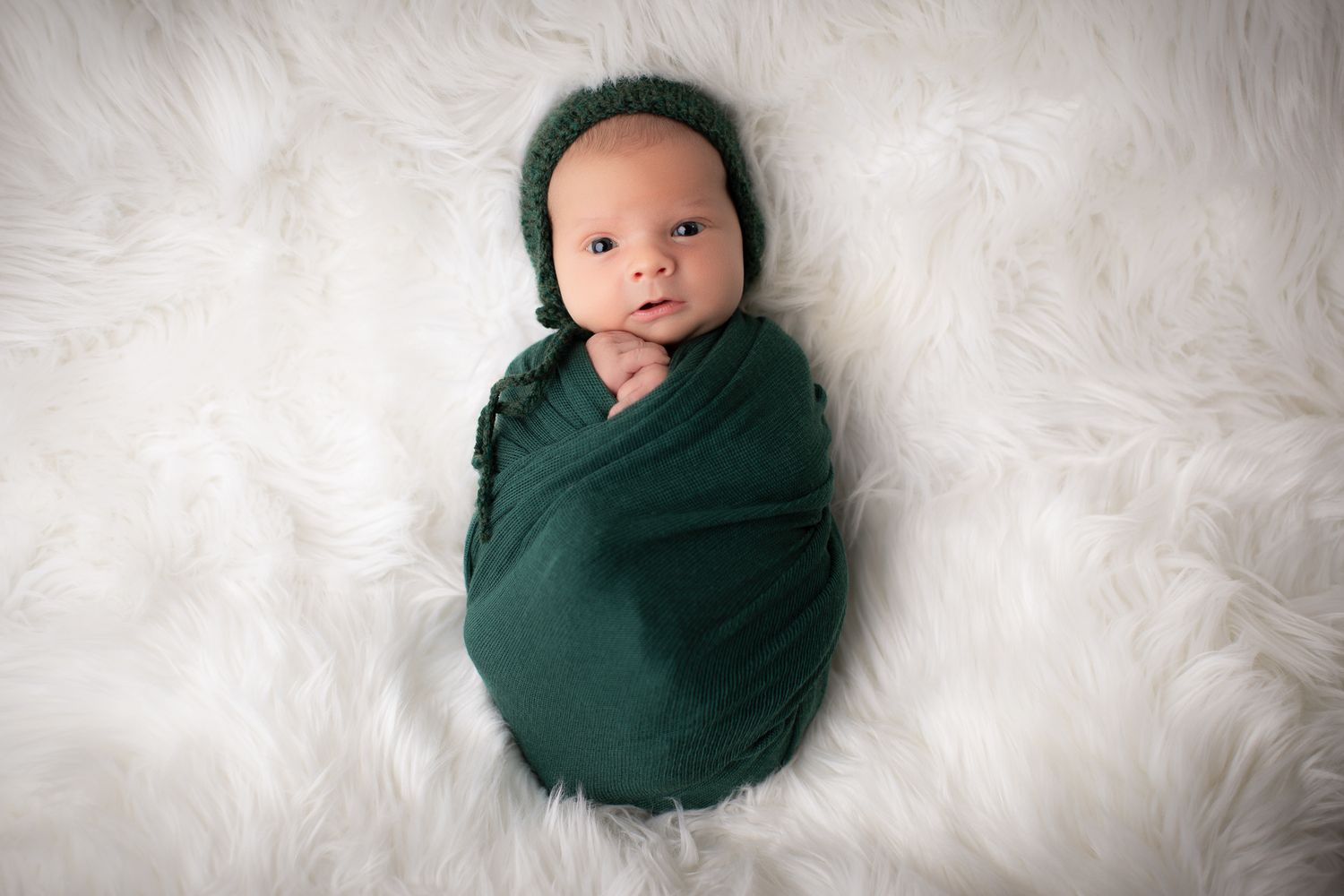 Newborn baby swaddled in dark green wrap and bonnet, lying on white fluffy fur backdrop.