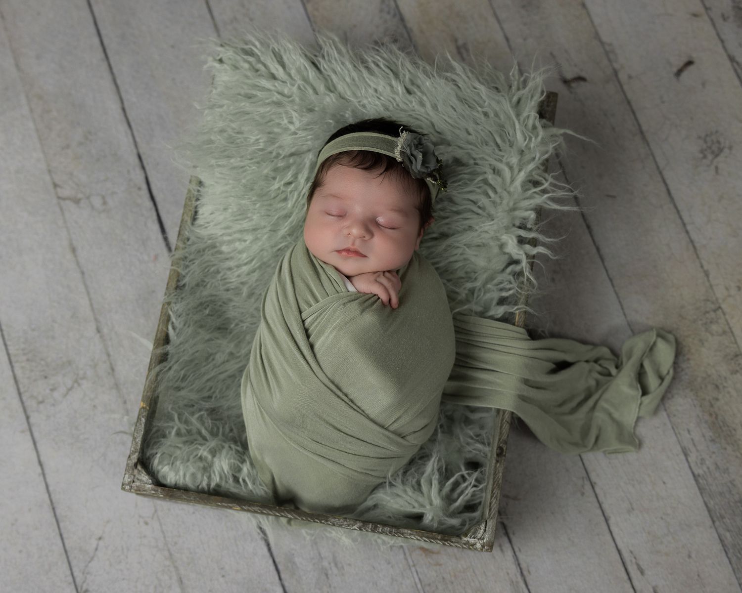 Sleeping newborn baby swaddled in sage green wrap with matching headband on fluffy green fur prop in San Marcos