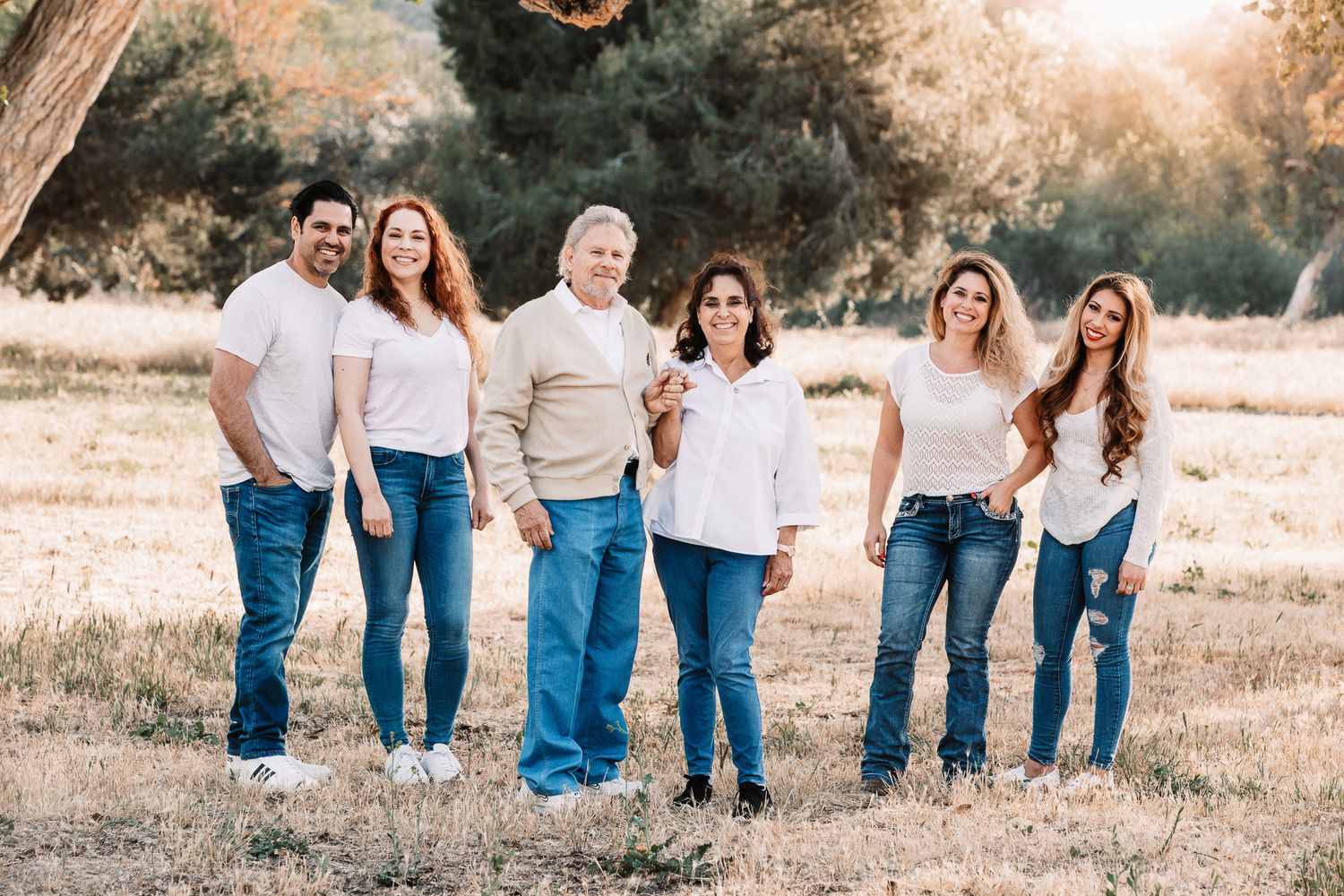 Extended family of six in matching white tops and jeans posing in golden meadow with trees.