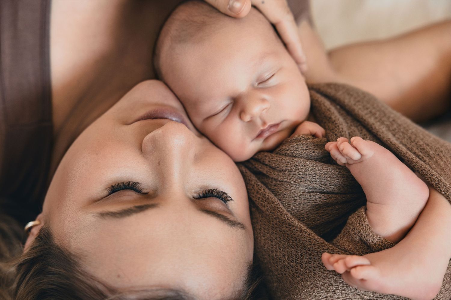 A mother tenderly rests her cheek against her sleeping newborn baby wrapped in a brown swaddle.
