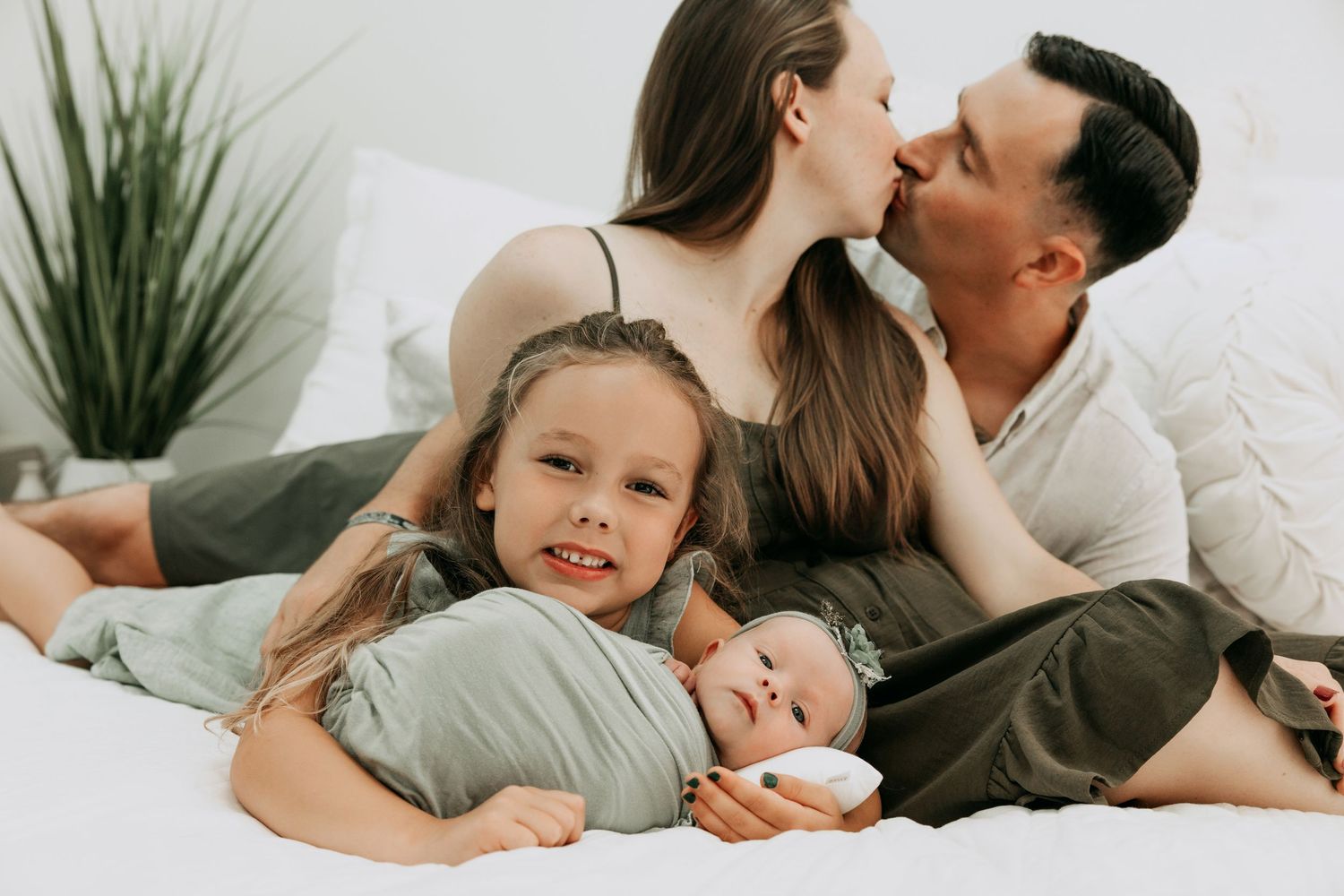 Happy family on white bed, parents kissing behind smiling girl holding newborn baby in cozy home setting.