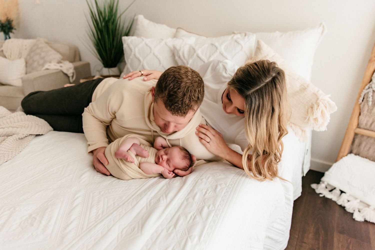 New parents lovingly gaze at their swaddled newborn baby on a white bed during an in-home newborn photography session.