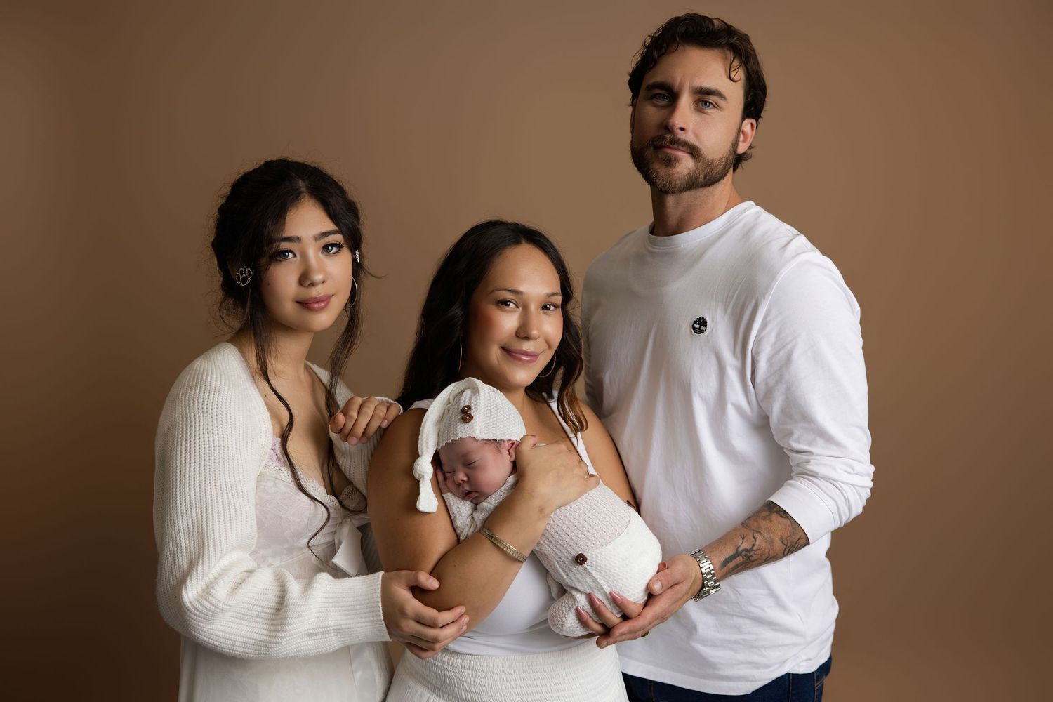Three people in white outfits pose for a newborn photo session, with one holding a sleeping baby in a white hat.