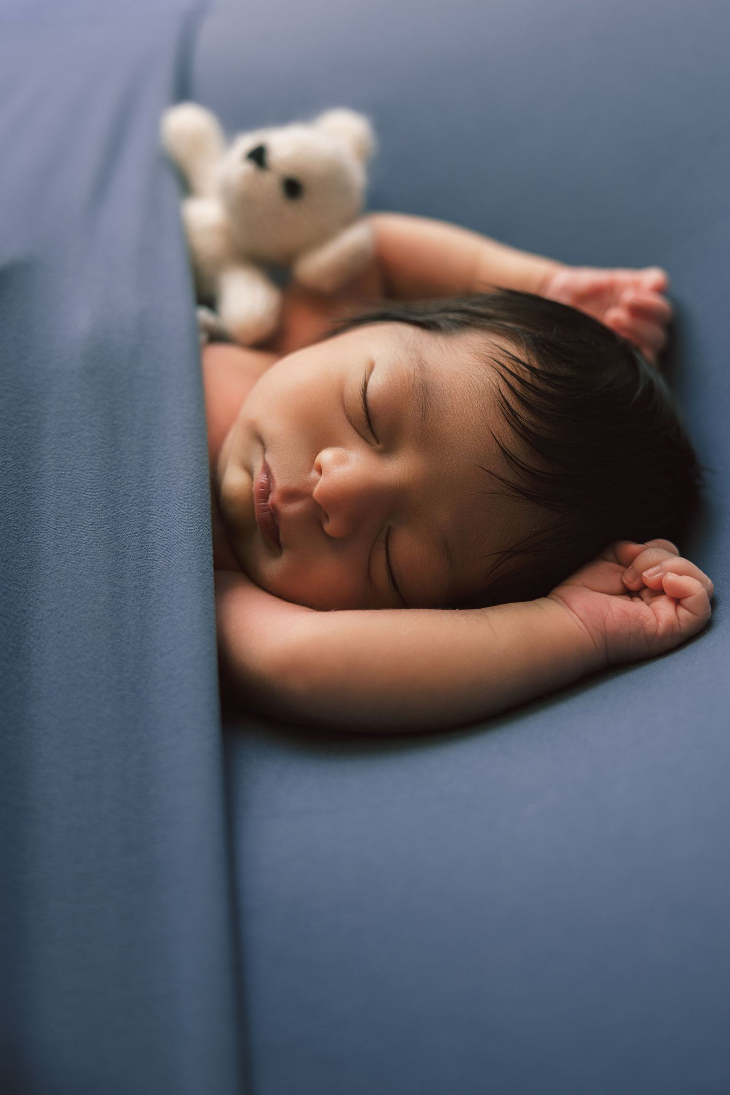 Newborn baby sleeping peacefully on blue surface beside a small white teddy bear.