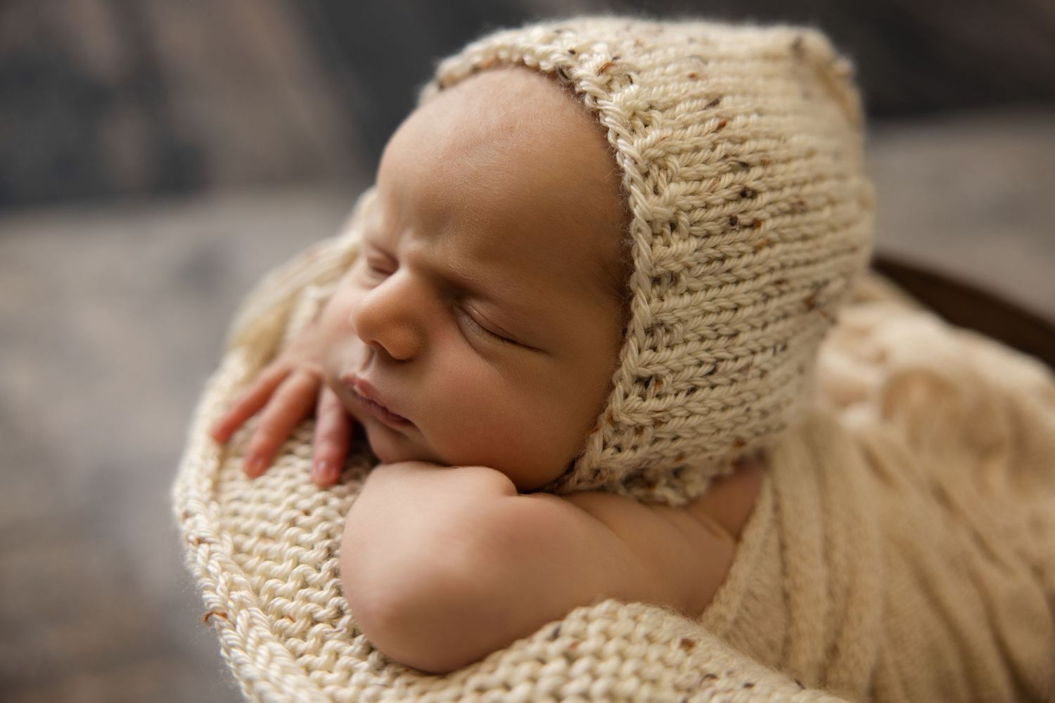 Peaceful newborn baby sleeping in a knit cream bonnet and wrap, nestled in a cozy bowl for a studio portrait.