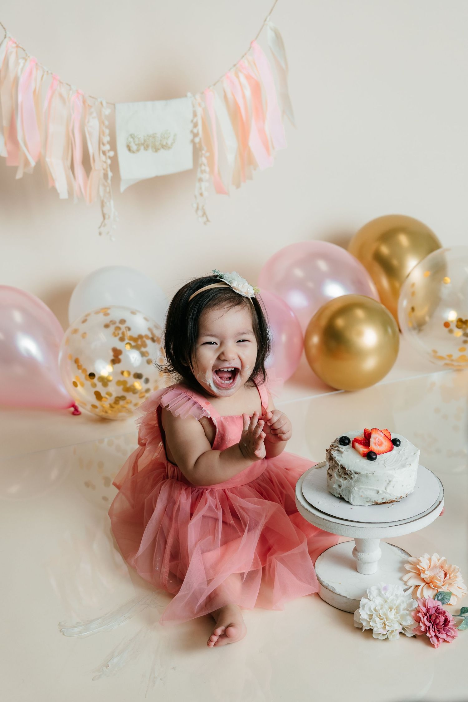 Happy baby girl in pink tutu laughing at first birthday cake smash with colorful balloons and ONE banner.