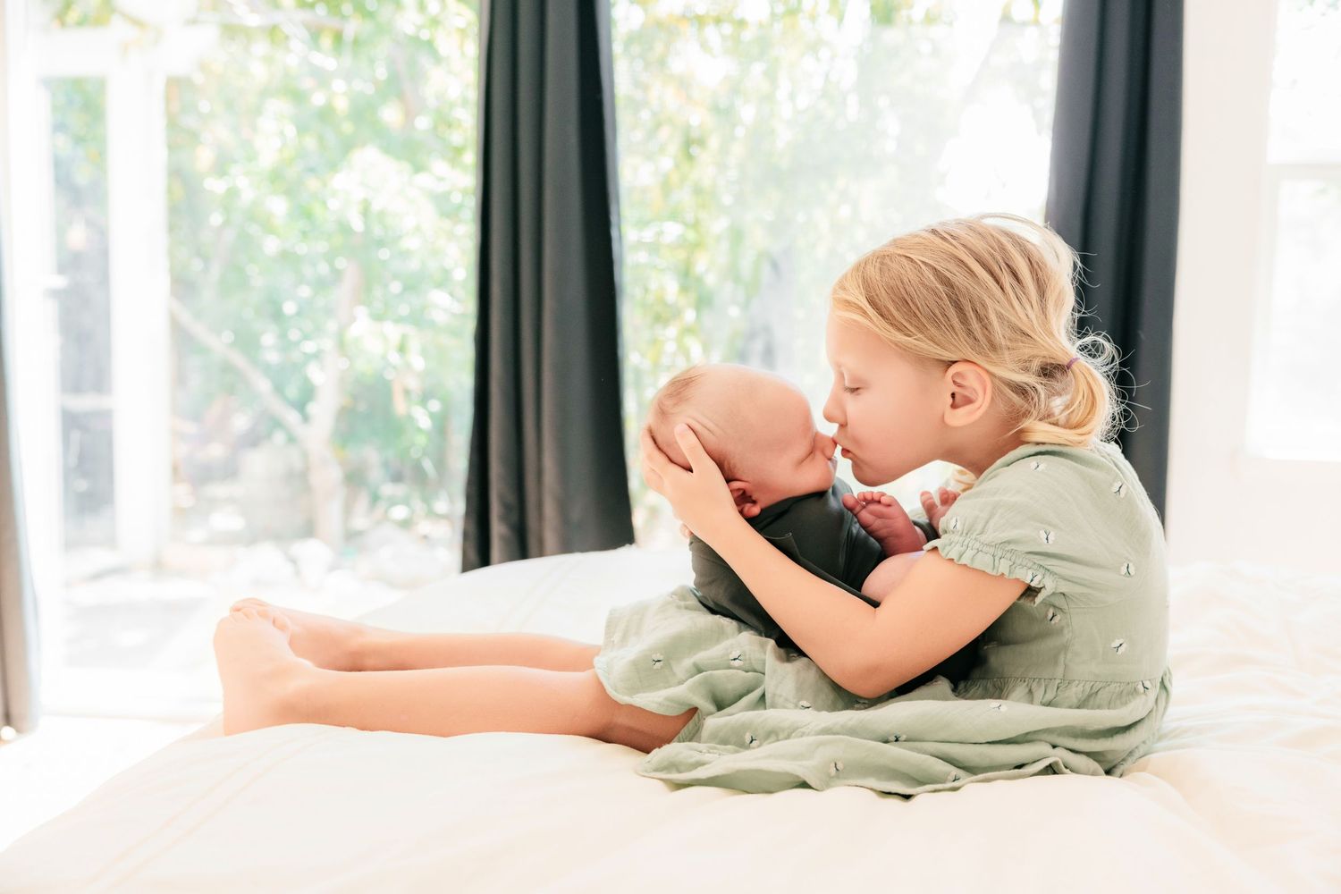 Young girl sitting on white bed gently kissing newborn baby sibling in a bright, light-filled room.