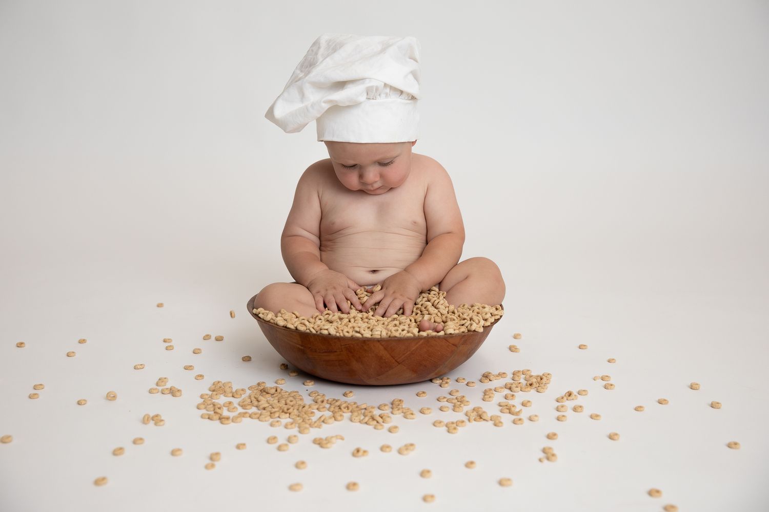 Cute baby wearing chef hat sitting with wooden bowl of cereal on white background.