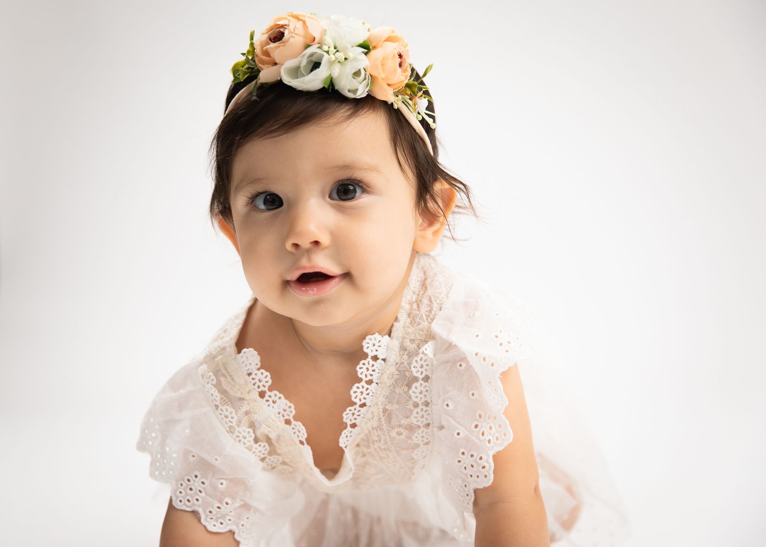 Beautiful baby girl wearing a floral crown and white lace dress looking at camera.