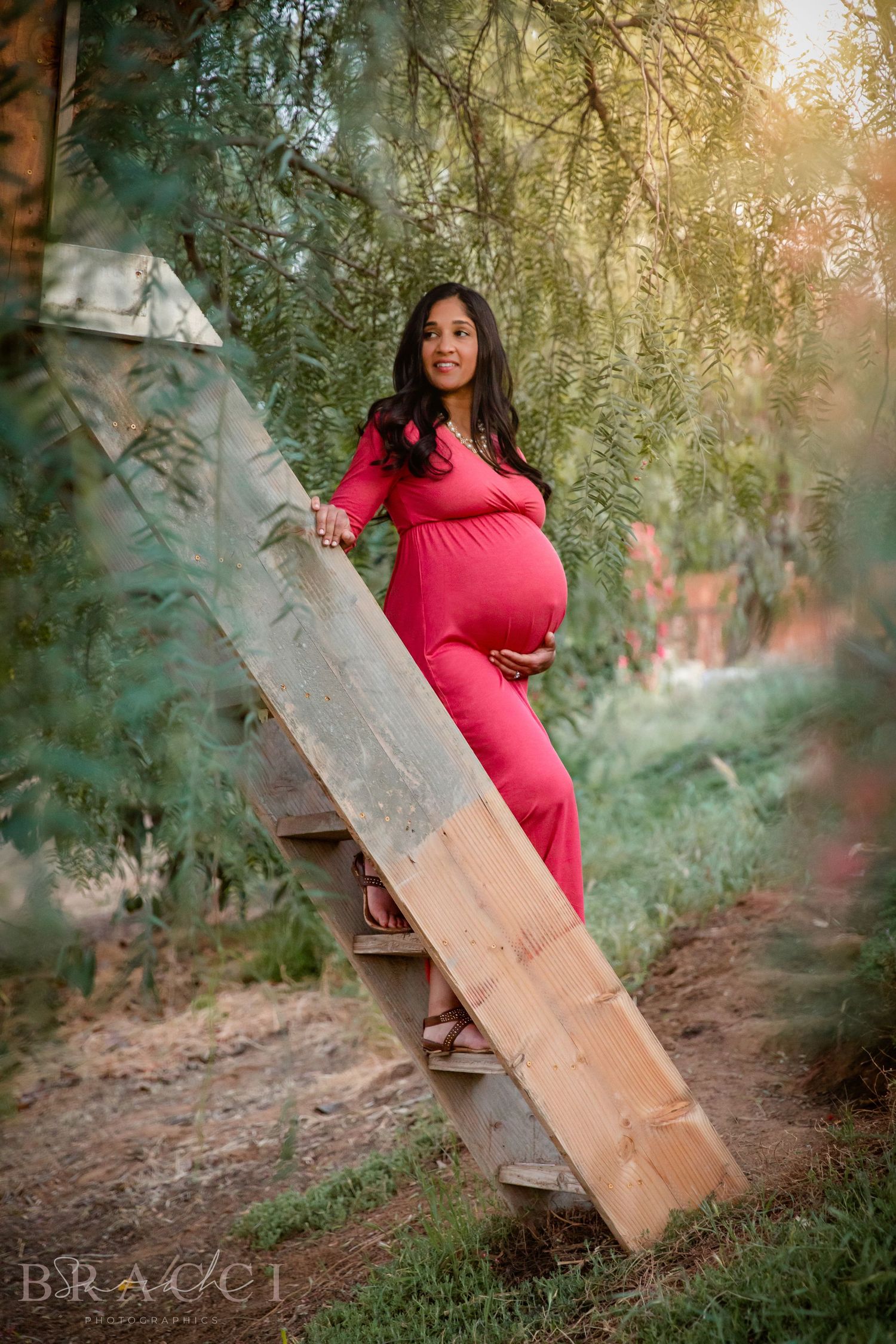 Smiling pregnant woman in a red maternity gown standing on wooden steps surrounded by lush green trees in Bonsall, near Fallbrook, & Temecul