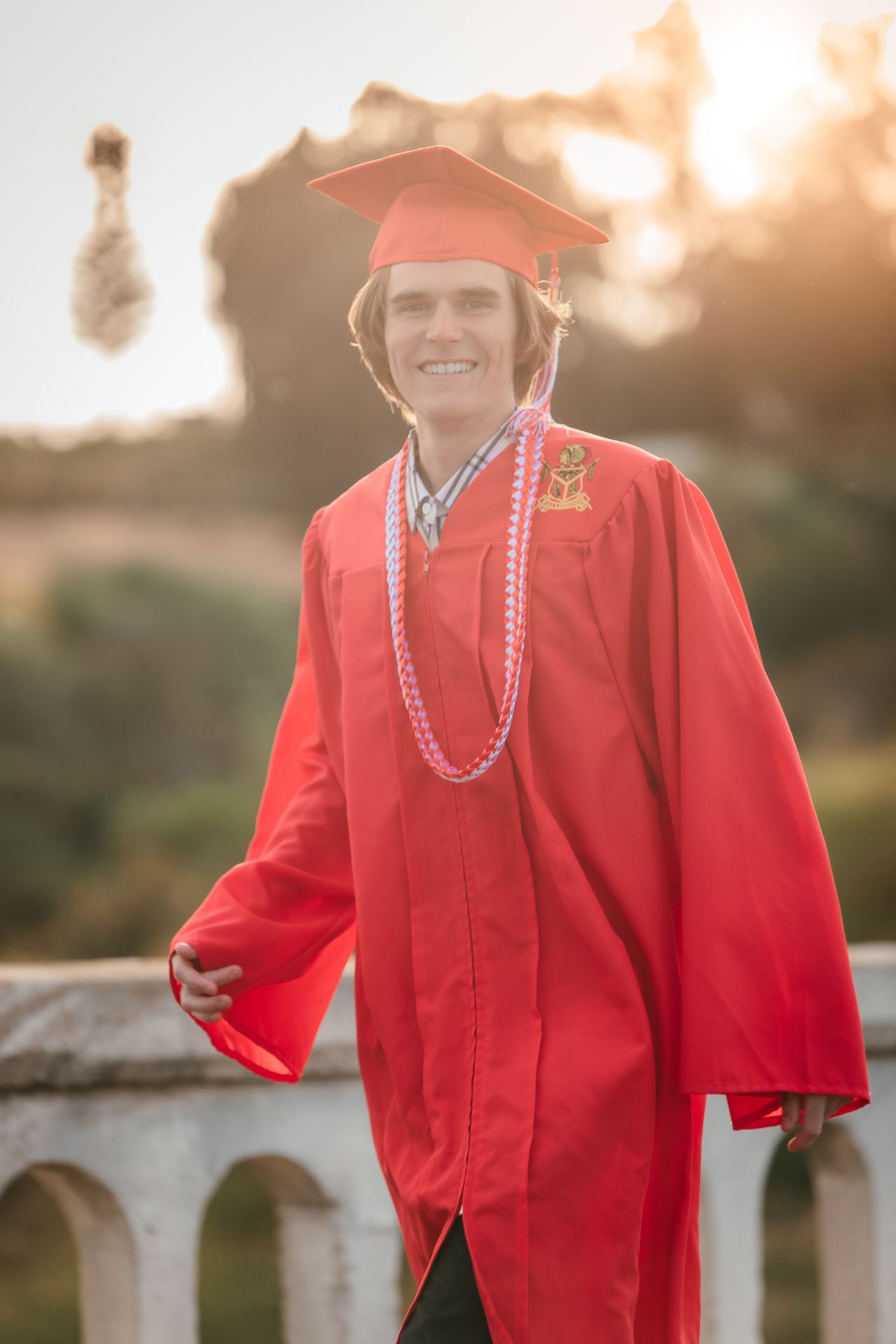 Smiling young man in red graduation cap and gown with honor cords posing outdoors in golden sunset light.