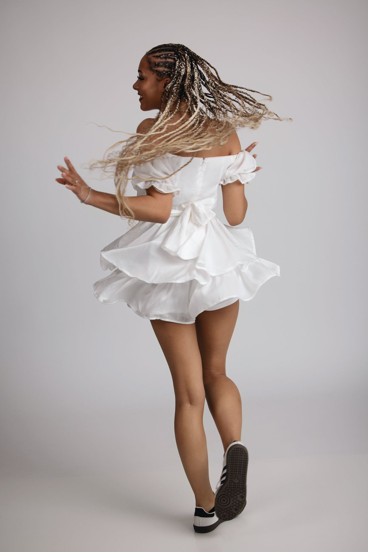 Young woman with long blonde braids spinning in white ruffled mini dress and sneakers in studio. Senior photography session in Bonasll near