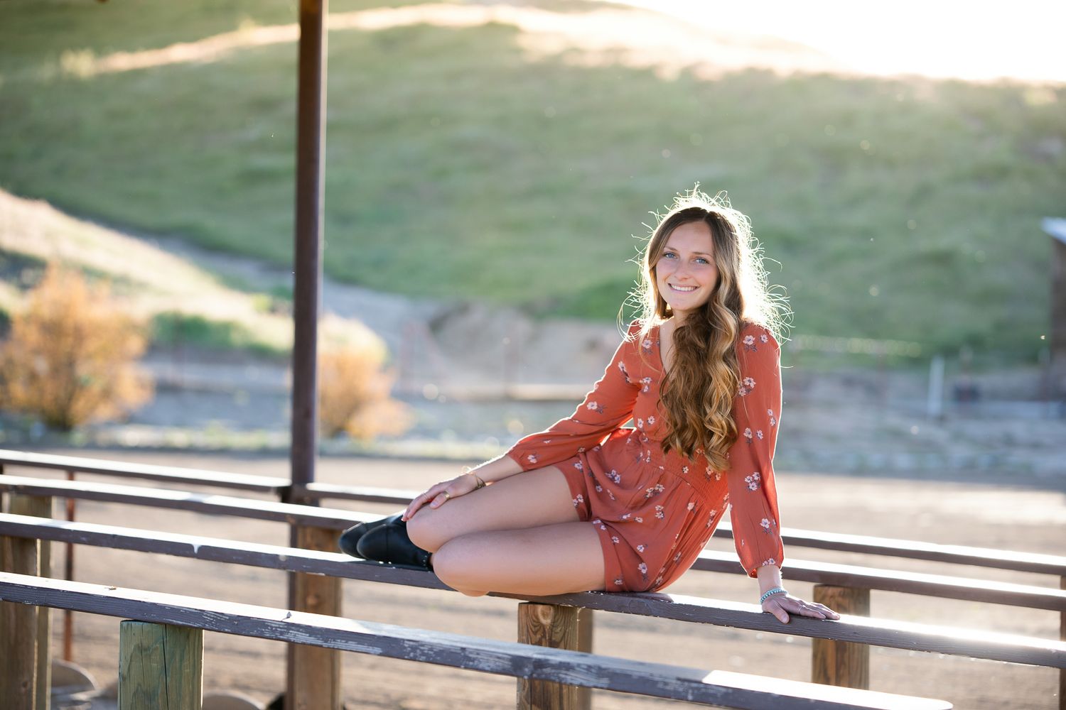 Smiling young woman in red floral dress sits on fence rails in sunny outdoor setting. Senior photography session in Temecula