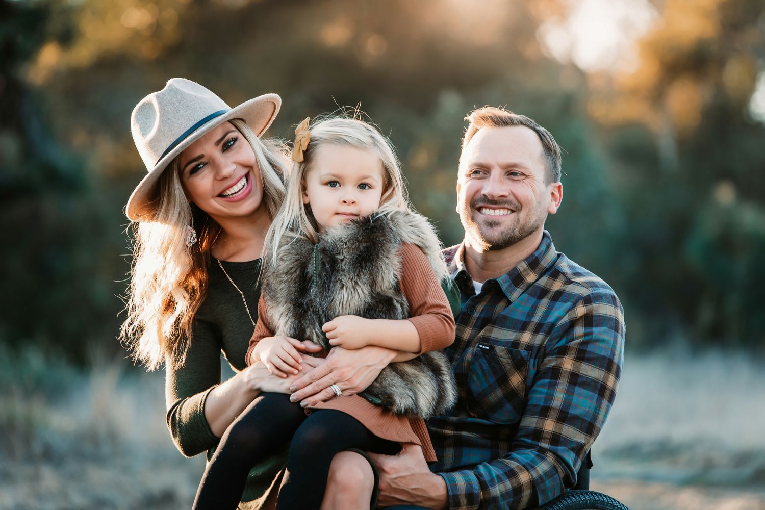 A smiling couple holds their young daughter outdoors in a warm, sunlit natural setting.
