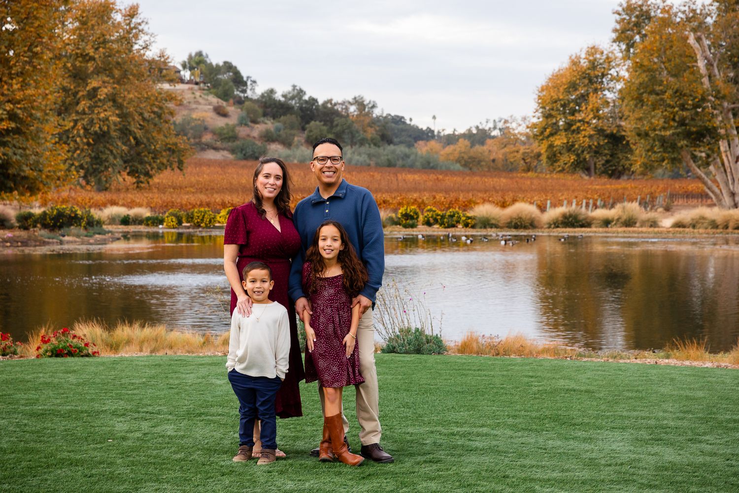 Family of four posing together near scenic pond surrounded by colorful fall foliage.