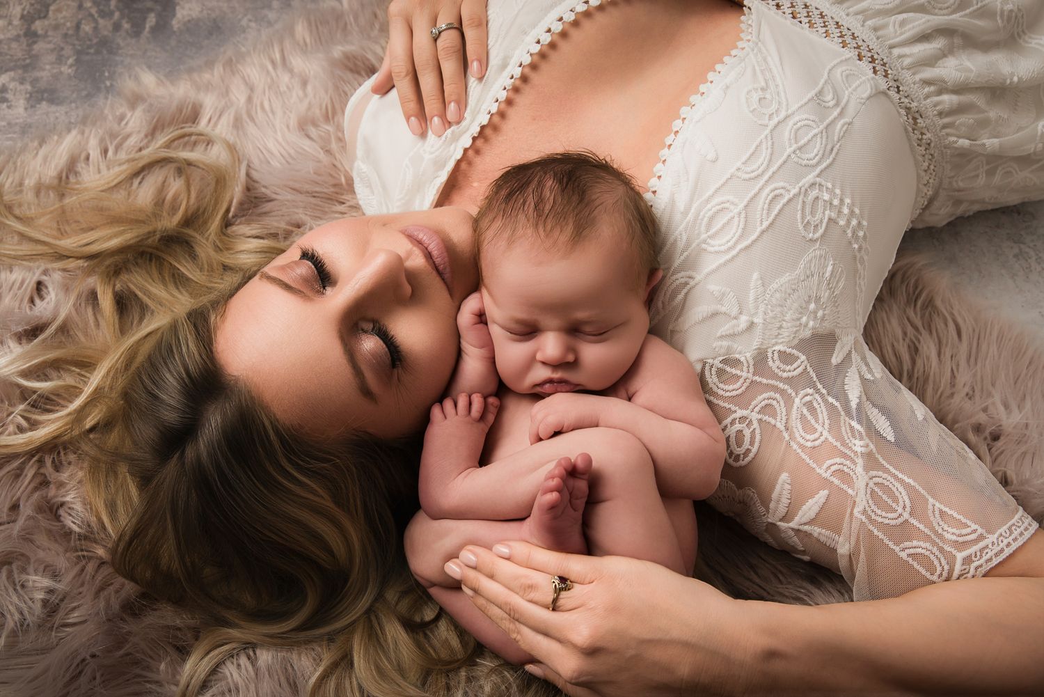 A mother in white lace dress lying down cheek-to-cheek with her sleeping newborn baby on a fluffy surface.