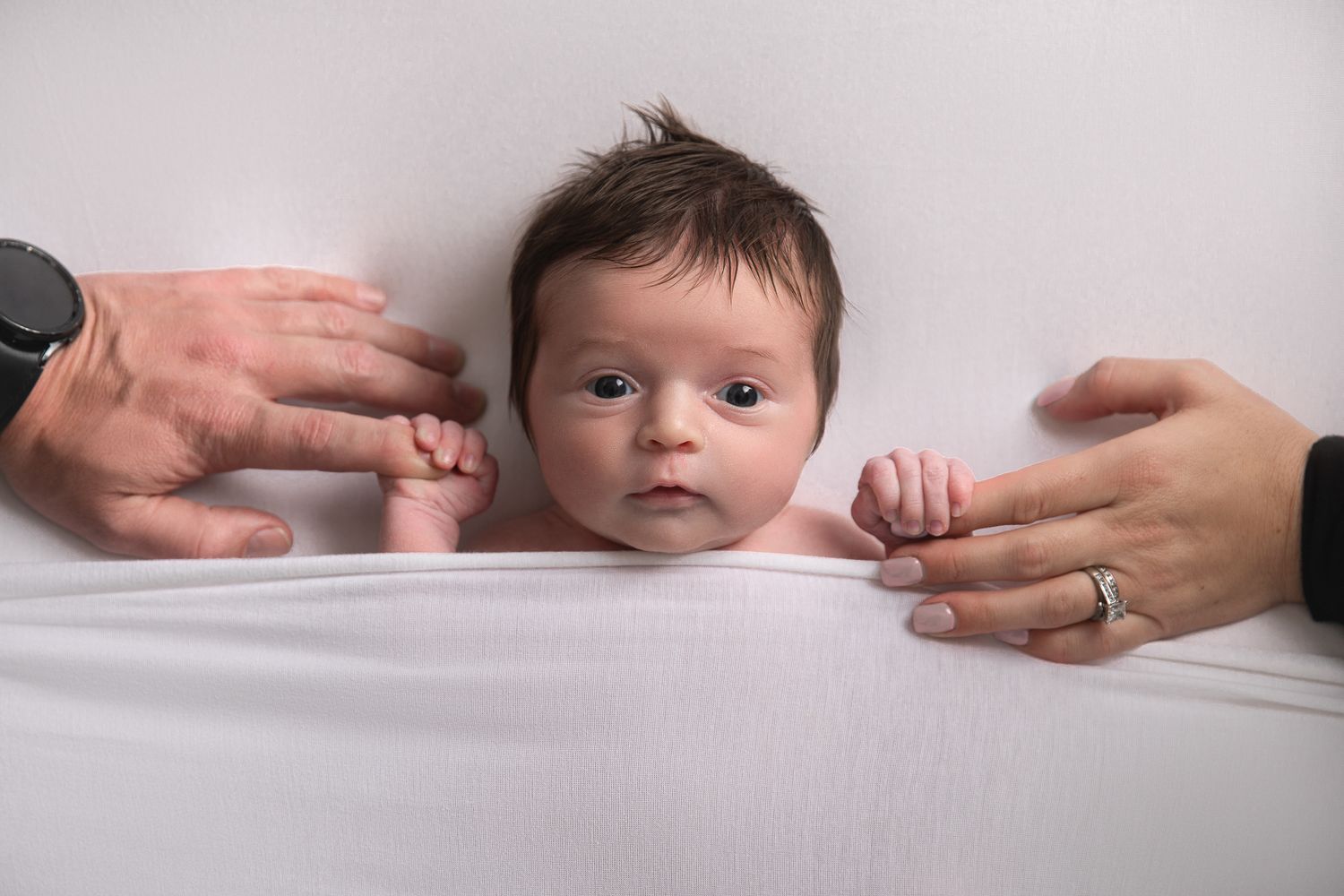 Alert newborn baby lying on white blanket with parents hands reaching in from both sides at The Studio Bonsall, near Vista & Oceanside
