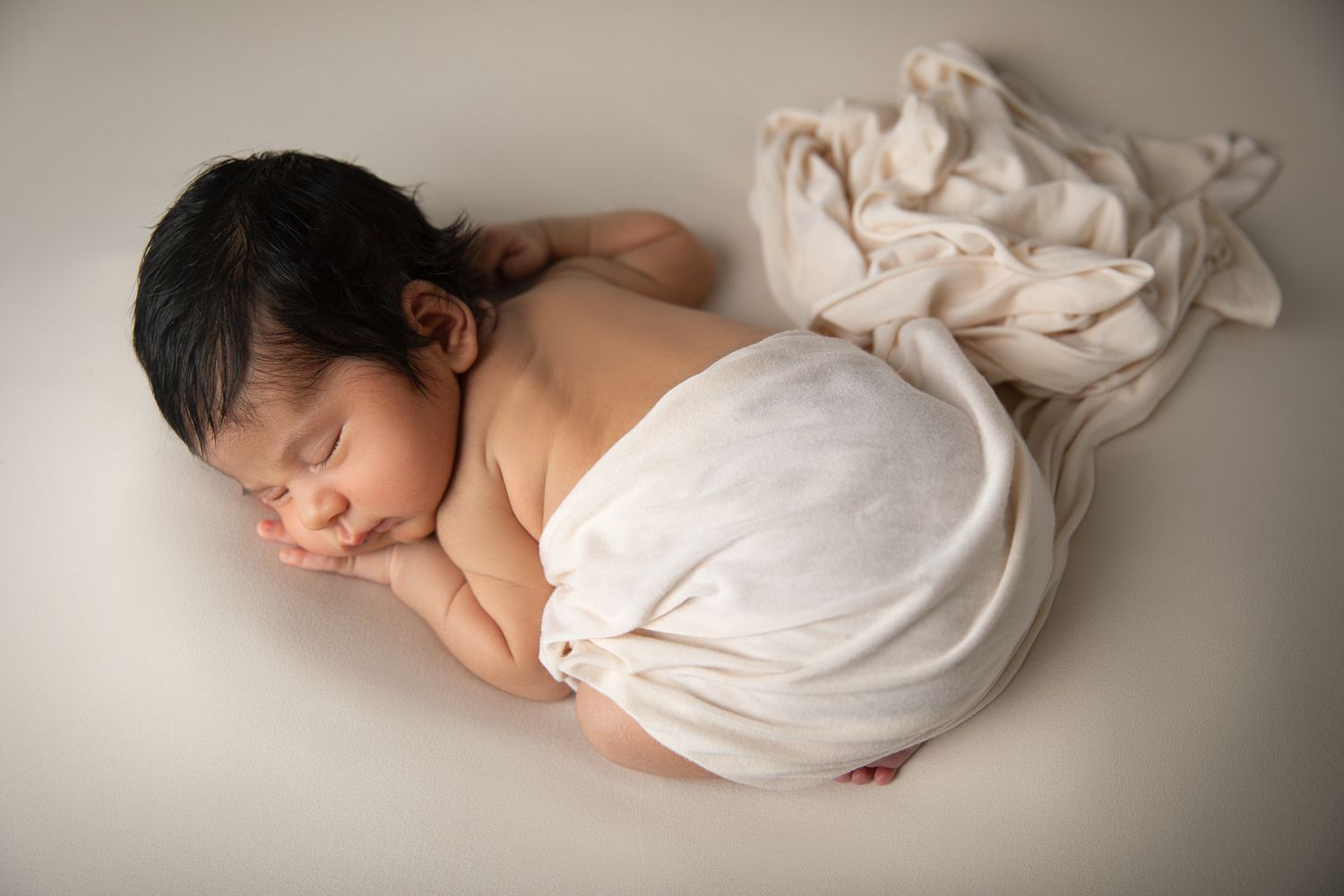 Sleeping newborn baby wrapped in white cloth posed on neutral background.