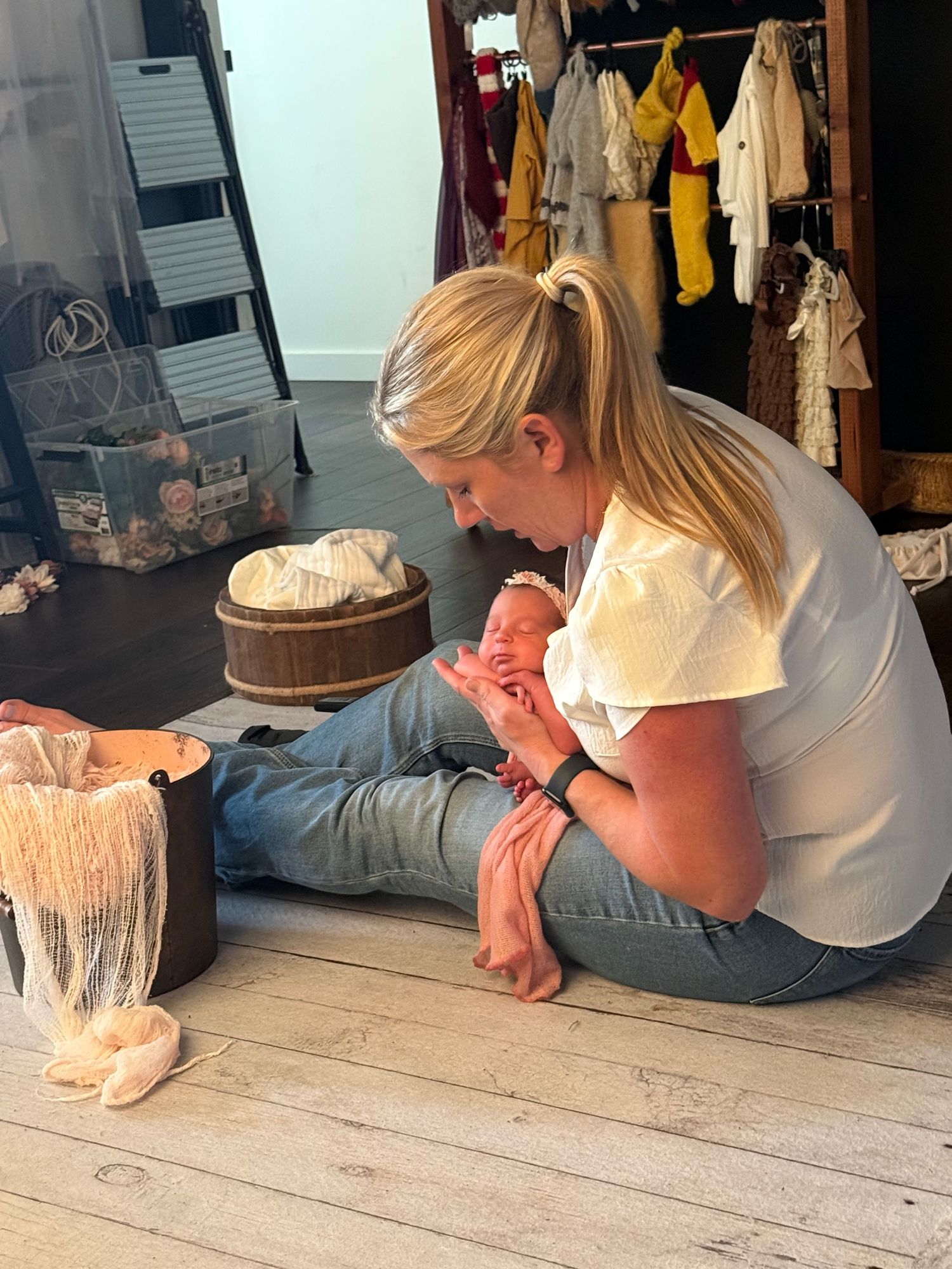 Blonde woman sitting on wooden floor holding a newborn baby during a professional photography session.