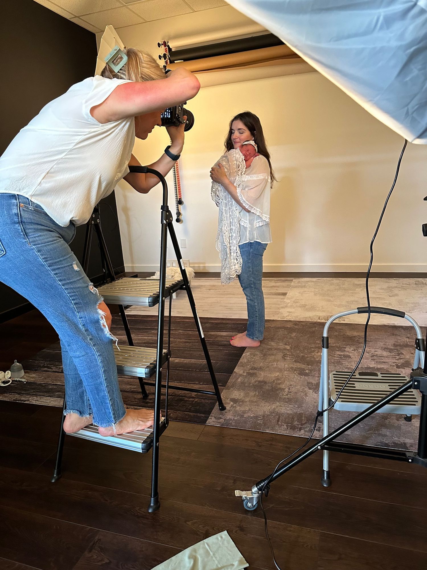 Photographer on stepstool shoots female model holding book in professional photography studio with lighting equipment.