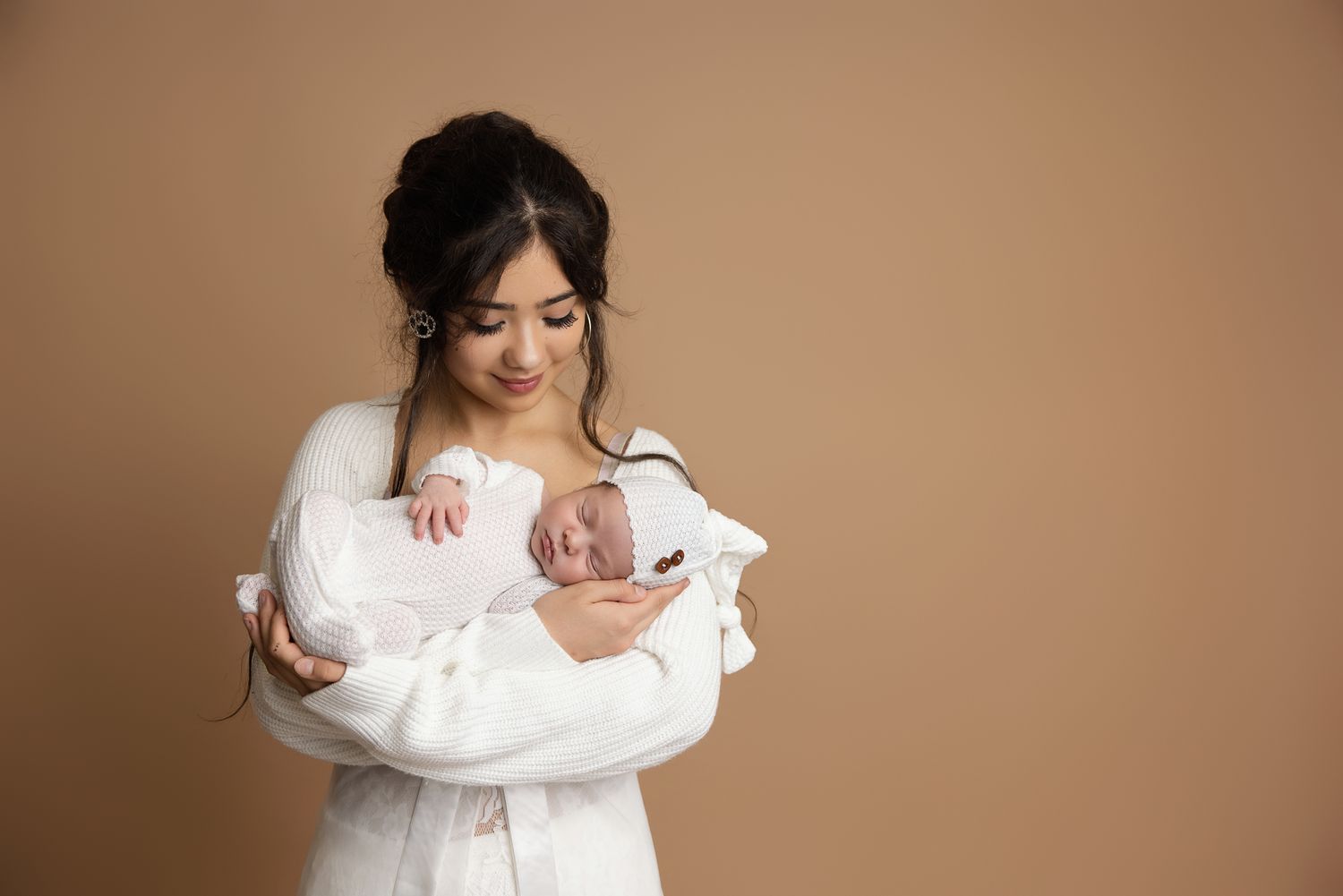 A young mother in a white robe tenderly holds her sleeping newborn baby against a warm beige background.