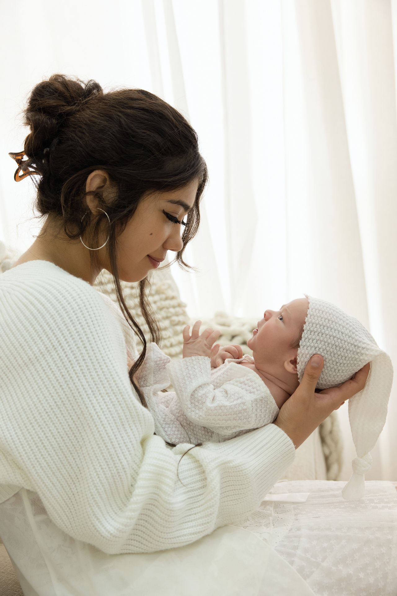 Young mother in white sweater tenderly holds her newborn baby wearing a white knit hat near a bright window.