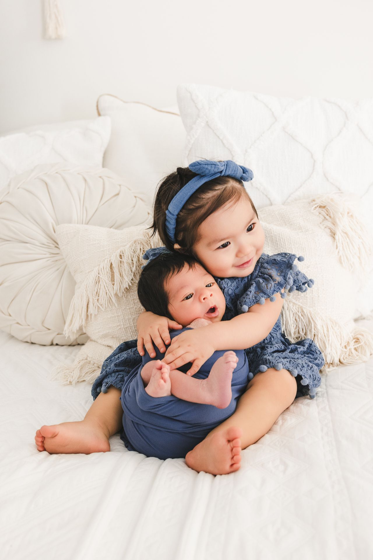 Young girl in blue dress hugs newborn sibling on white bed with decorative pillows.