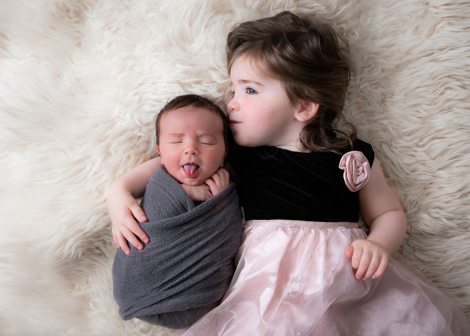 Newborn baby swaddled in gray wrap yawning beside older sister in black velvet and pink tutu dress on fluffy white rug.