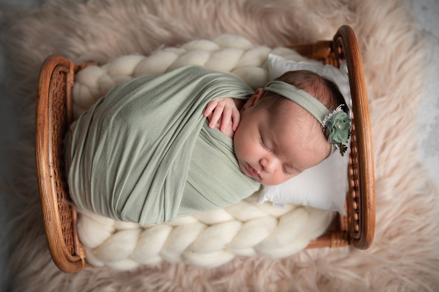 Sleeping newborn baby girl swaddled in sage green wrap with floral headband in a wooden bed.