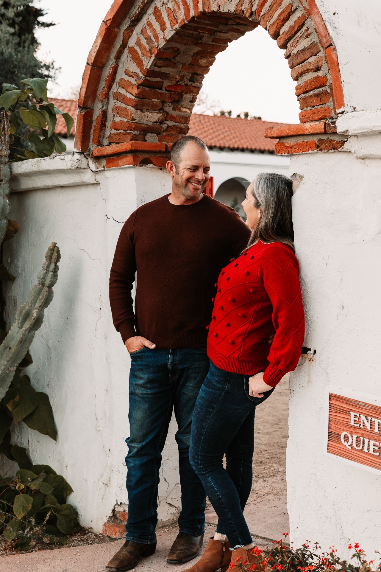 Couple sharing a smile under brick arch at historic outdoor venue in cozy sweatersin Bonsall, near Fallbrook, & Temecula