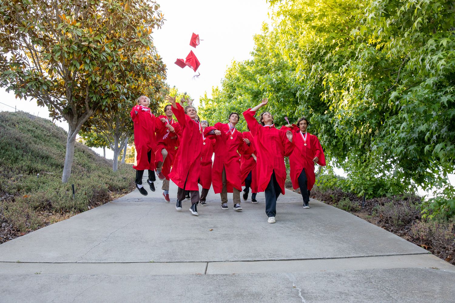 Group of graduates in red caps and gowns celebrating outdoors on pathway.