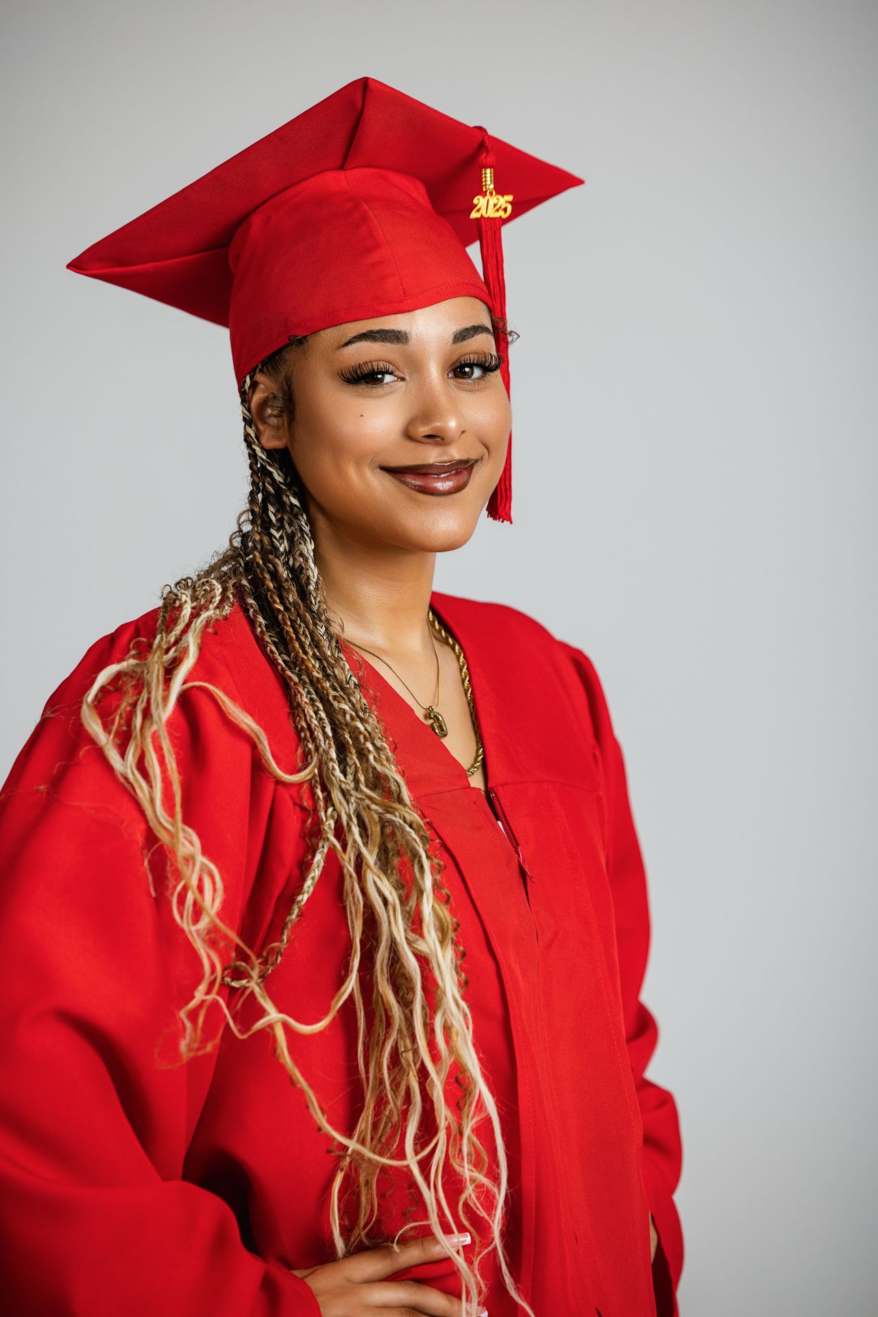 Smiling graduate in red cap and gown with long braids poses for a 2025 graduation portrait. Senior photography session in the studio bonsall