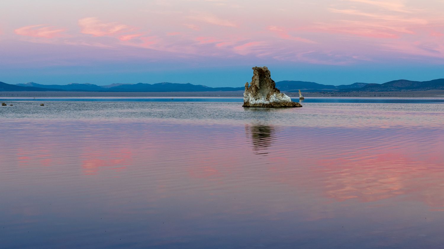 A lone tufa rock formation reflects in the calm waters of Mono Lake at sunset with pink and blue skies.