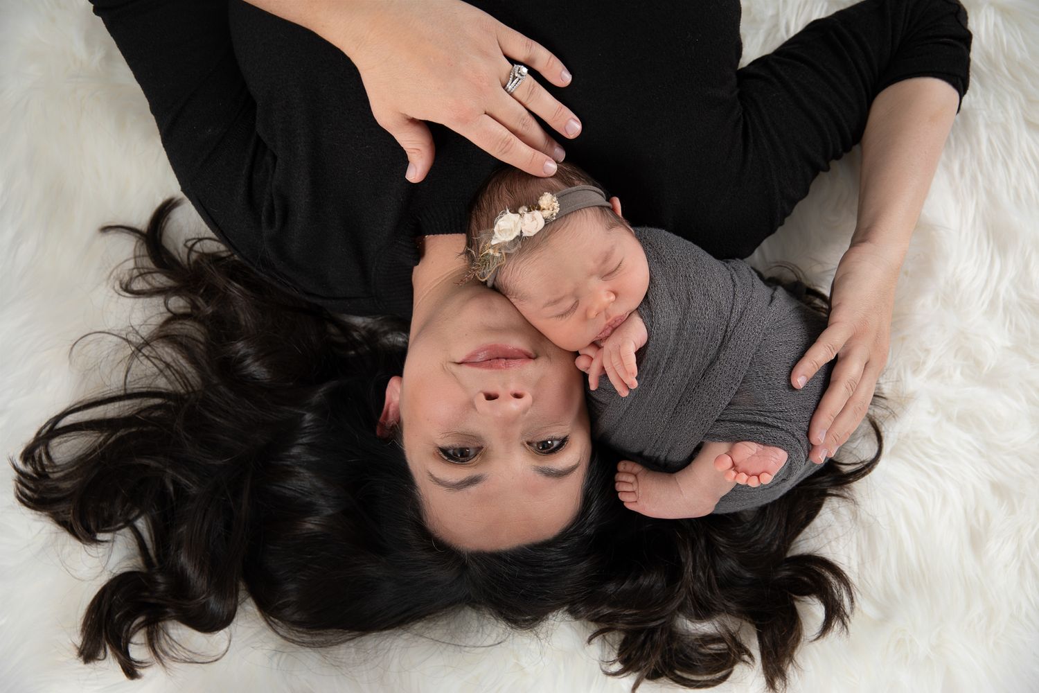 A mother with dark hair lies on a white fur rug with her sleeping newborn baby wearing a floral headband.
