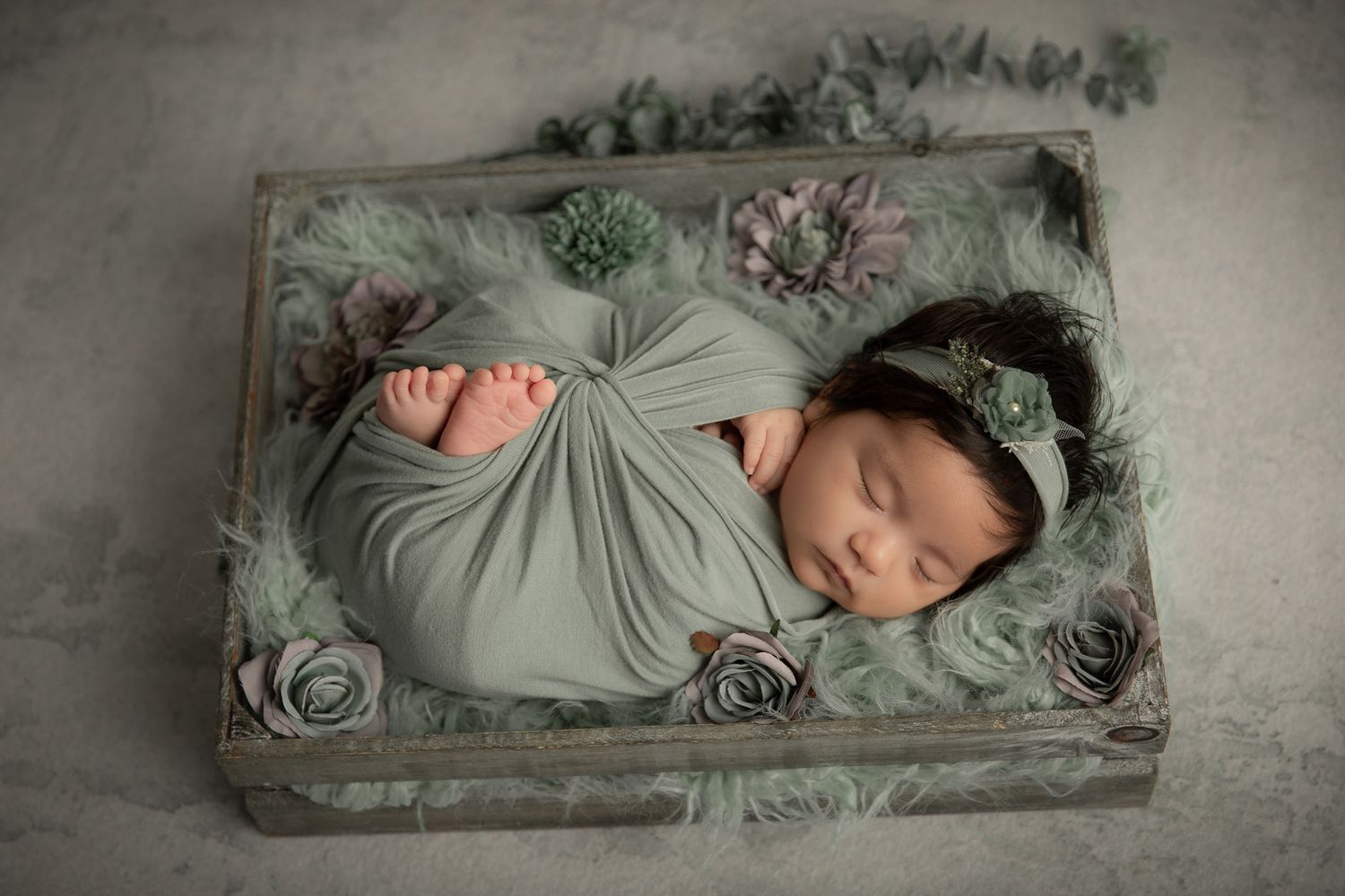 Newborn baby wrapped in sage green swaddle sleeping in a wooden box with floral decorations and a green headband.