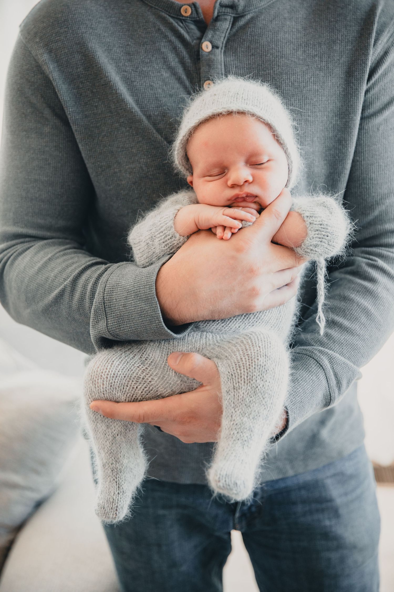 Father cradling sleeping newborn baby dressed in soft gray knit outfit and bonnet hat.