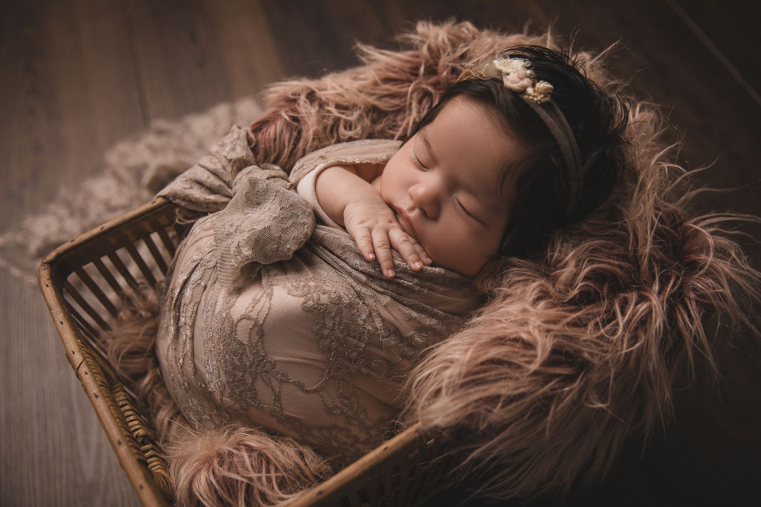 Newborn baby sleeping peacefully in a wicker basket with fluffy fur, wrapped in a soft brown swaddle with a floral headband.