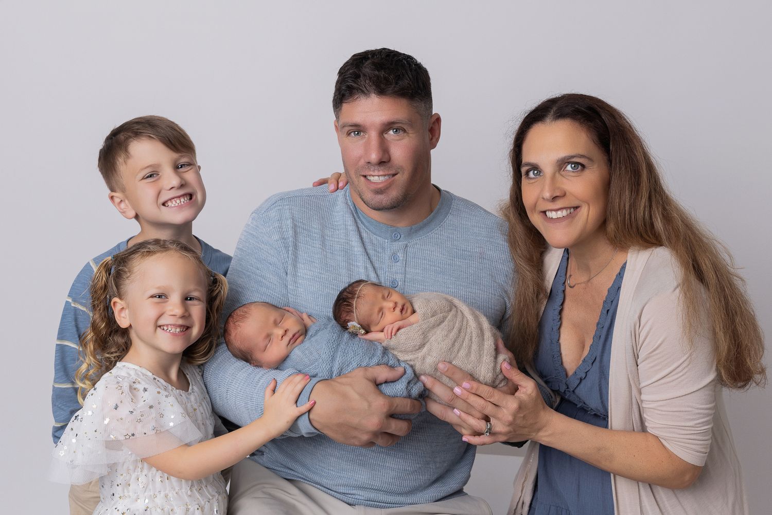 Family of five posing for newborn portrait with parents holding twin babies and two older siblings smiling.