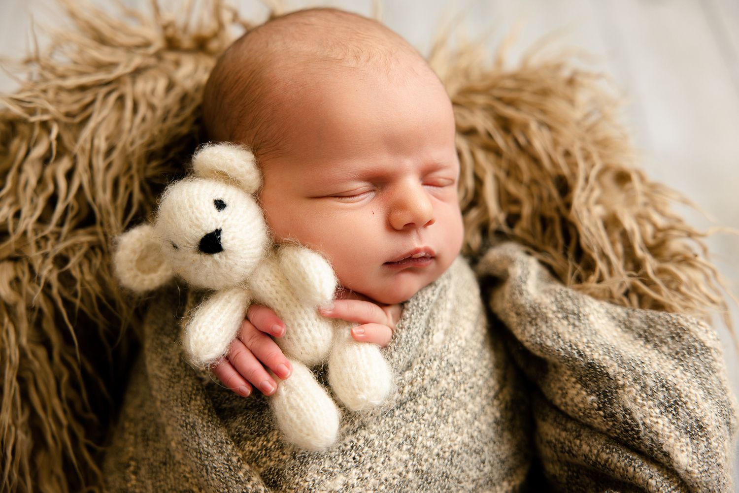 Newborn baby sleeping peacefully in a basket wrapped in a knit blanket holding a small white teddy bear.