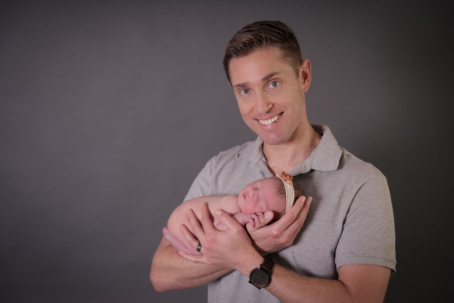Smiling man holding a sleeping newborn baby against a gray studio background.