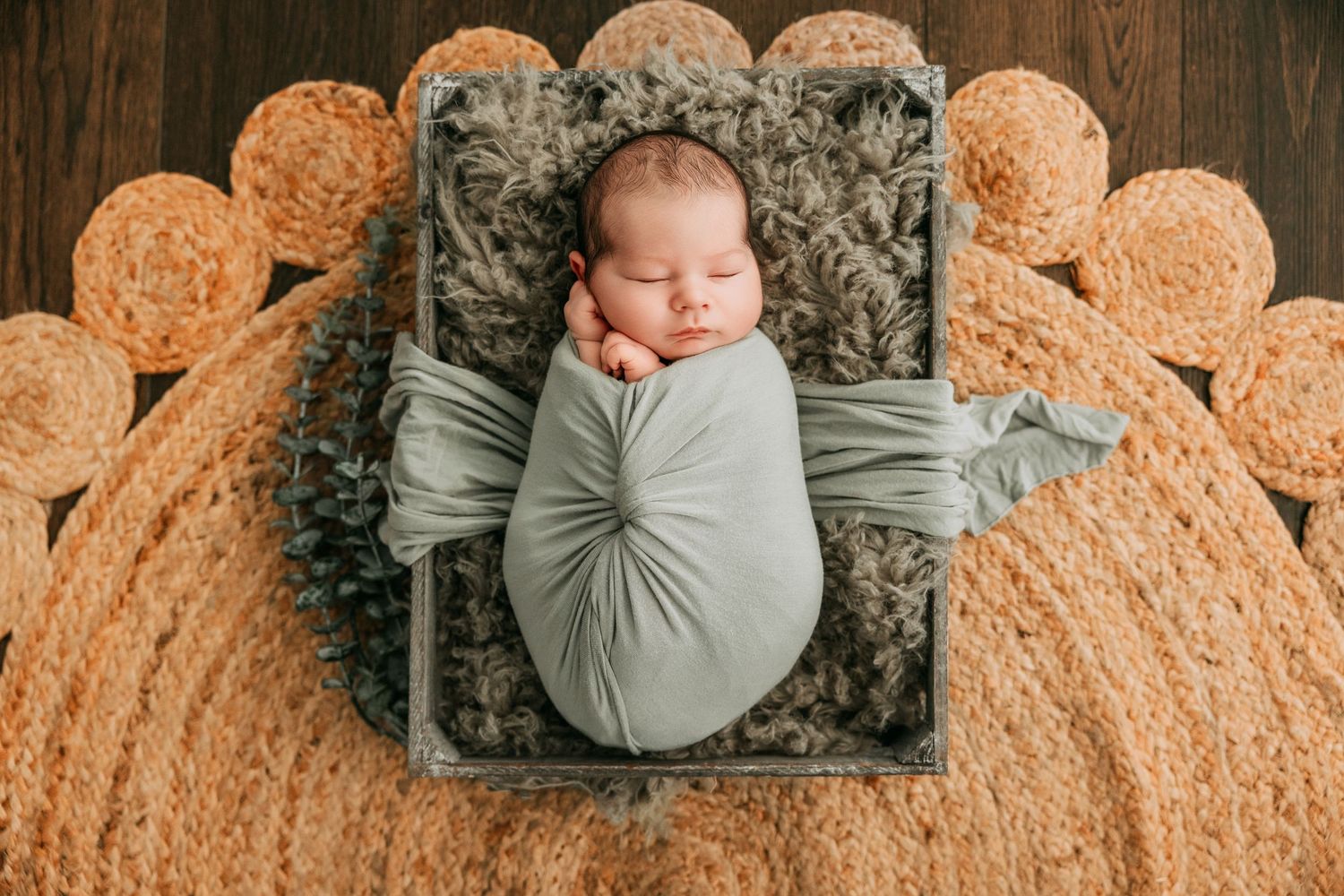 Newborn baby wrapped in sage green swaddle resting in a wooden crate on a textured sunflower rug.