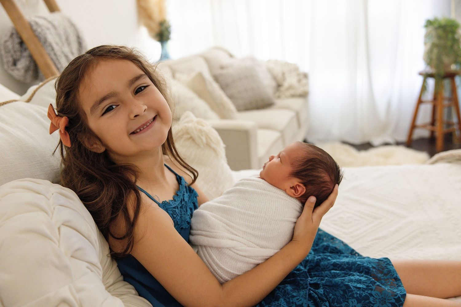 Young girl smiling while gently holding a swaddled newborn baby on a white couch indoors.