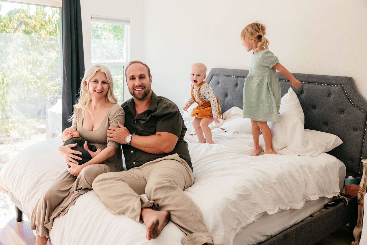 A happy family with two young children playing on a bed in a bright white bedroom, parents smiling at camera.