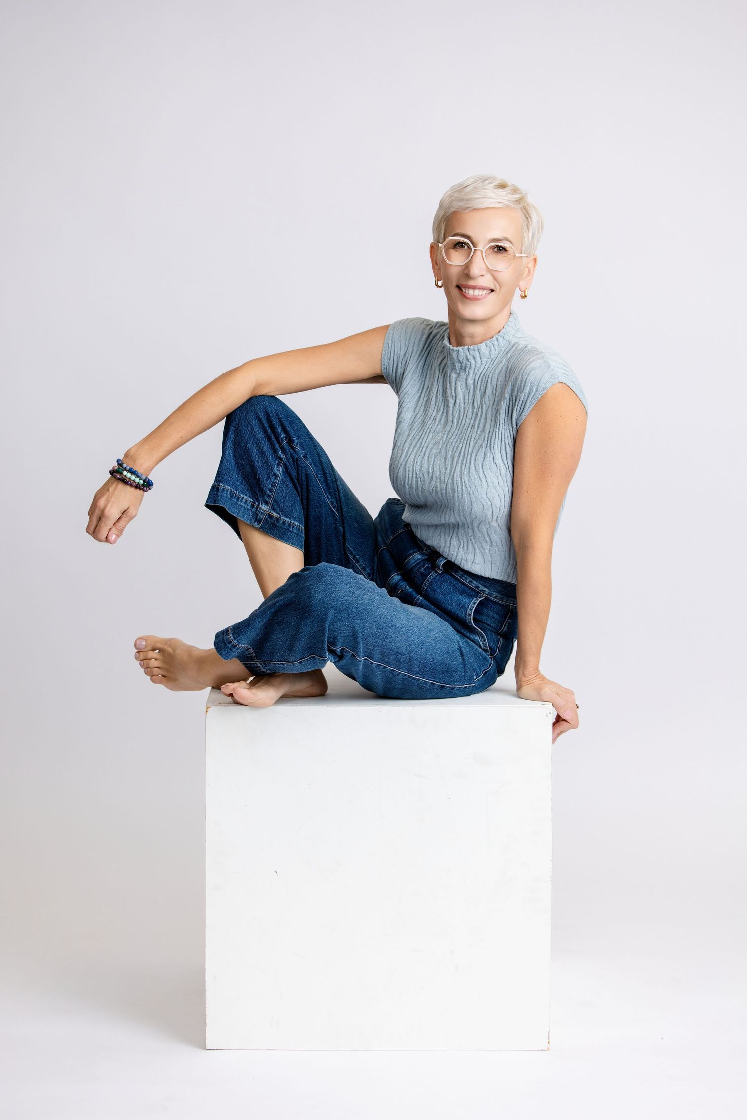 Smiling middle-aged woman with short gray hair and glasses sitting cross-legged on white cube in jeans and blue top.