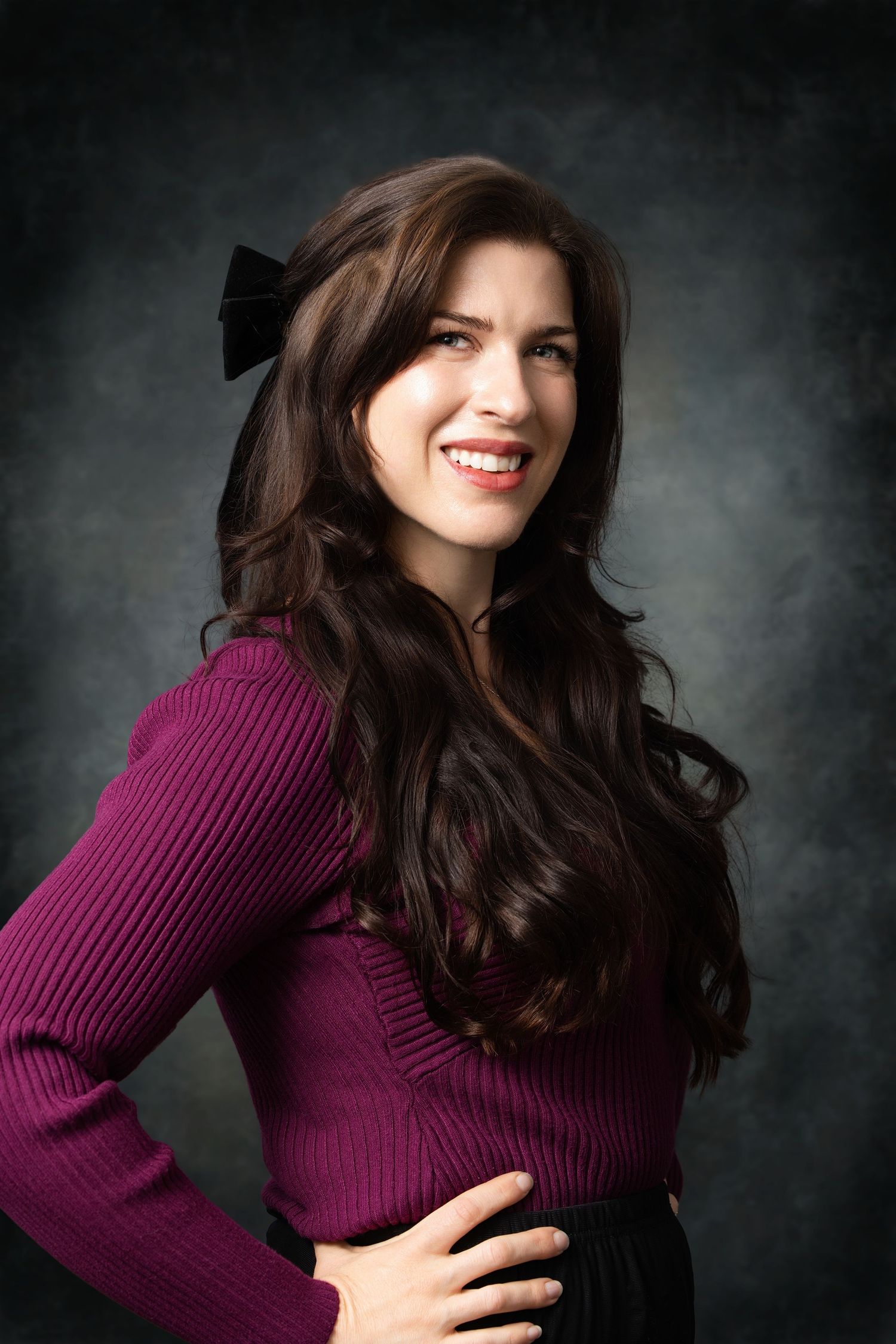 Smiling brunette woman with long wavy hair and black bow, wearing a purple ribbed sweater, posing against dark background.