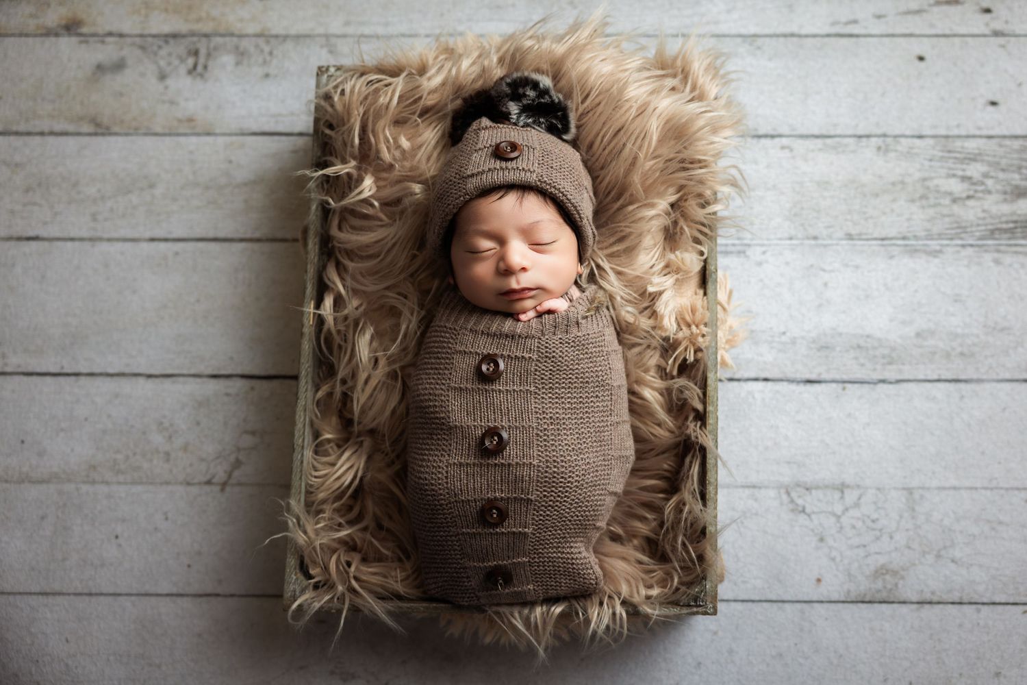 Newborn baby swaddled in brown knit wrap and hat, sleeping peacefully on fluffy fur in a rustic wooden box.