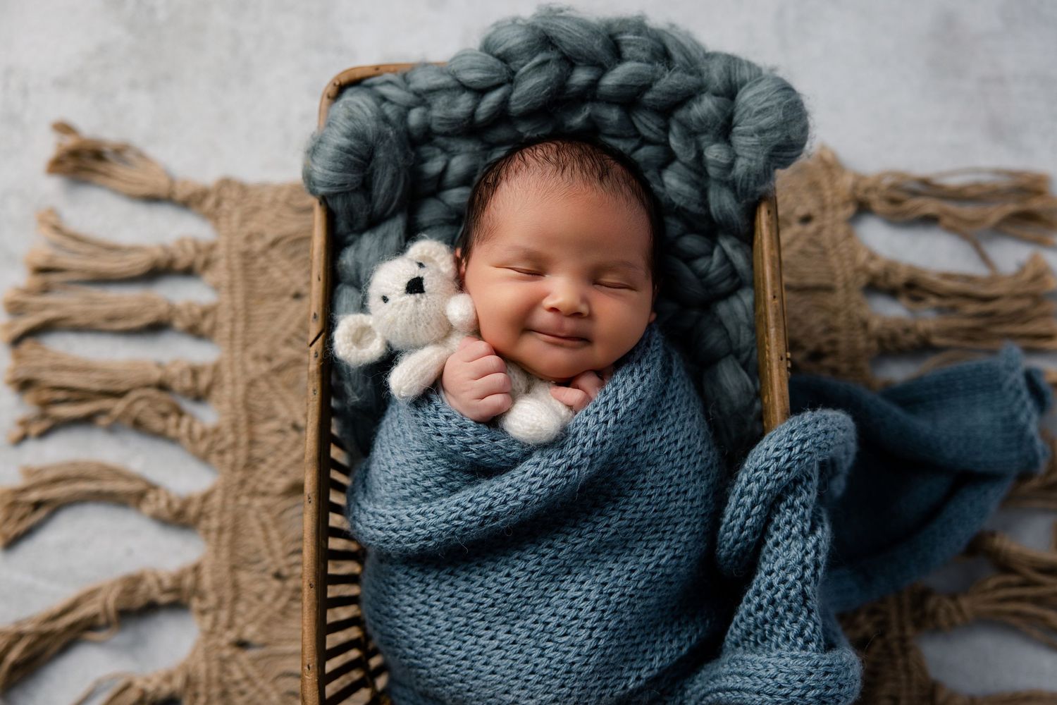 Smiling newborn baby wrapped in blue knit blanket holding teddy bear in basket with chunky yarn backdrop.
